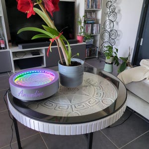May include: A round kinetic sand table with a glass top featuring a white sand pattern, accompanied by a circular light-up device with a purple frame and the name Amandine in script, and a gray pot containing a red amaryllis flower plant.