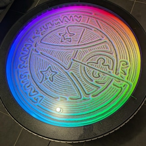 Op de afbeelding: Een ronde kinetische zandtafel met een zwart frame en een levendige regenboog LED-ring. Het witte zandoppervlak toont een complex, ingegraveerd geometrisch patroon met sterren en gebogen lijnen, gevormd door een metalen bal in het zand.