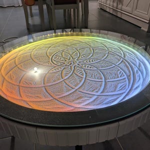 Peut inclure: Table basse ronde avec plateau en verre et motif de sable. Le motif est éclairé par un dégradé de couleurs allant du jaune à l'orange et au bleu. La table a un socle gris foncé.