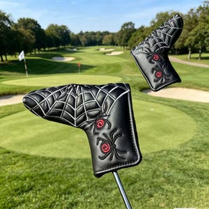 Könnte beinhalten: Zwei schwarze Golf-Putter-Abdeckungen mit weiß gestickten Spinnennetzmustern und zwei Spinnen mit roten kreisförmigen Zentren. Die Abdeckungen bestehen aus einem glatten Material und dienen zum Schutz des Kopfes eines Golf-Putters.