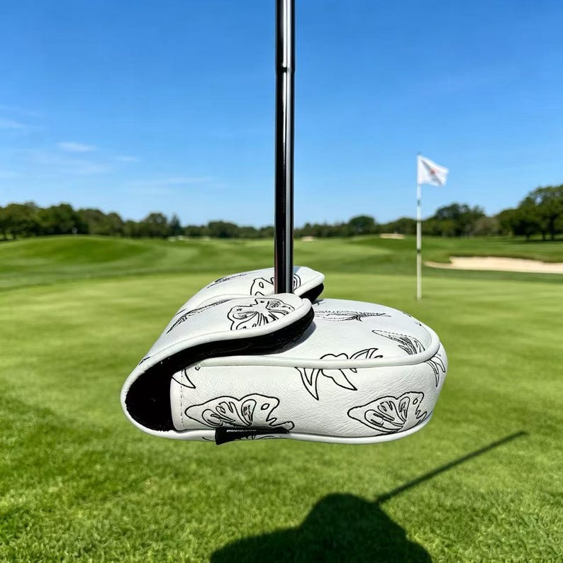 May include: A white golf putter headcover featuring a black line art butterfly pattern. The cover is constructed from a smooth material with a black interior lining and is attached to a metallic silver putter shaft, positioned against a golf course.