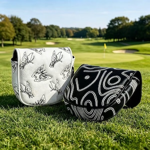May include: Two golf putter headcovers made of synthetic leather. One cover is white with a black butterfly pattern, and the other is black with a white abstract topographic line design. Both covers feature a structured, rounded shape.