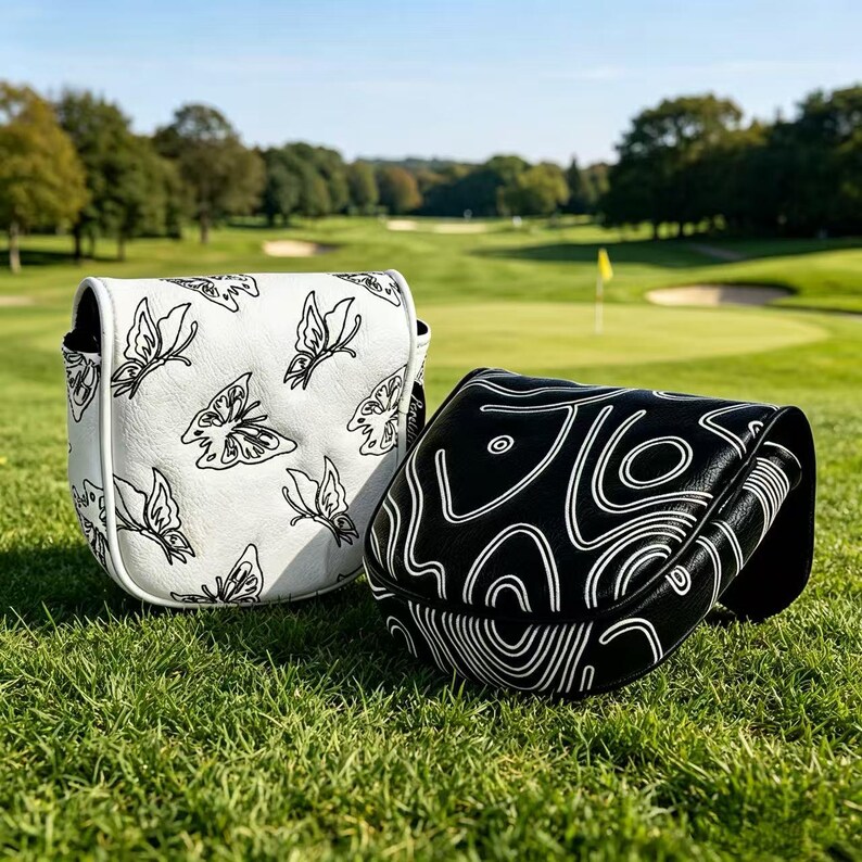May include: Two golf putter headcovers made of synthetic leather. One cover is white with a black butterfly pattern, and the other is black with a white abstract topographic line design. Both covers feature a structured, rounded shape.