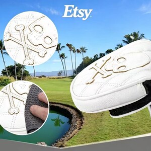 Puede incluir: Una funda blanca para putter de golf con textura acolchada y una aplicaci&oacute;n de color tostado en forma de calavera y tibias cruzadas. La funda tiene un forro interior negro y est&aacute; unida a un eje de putter de metal plateado.