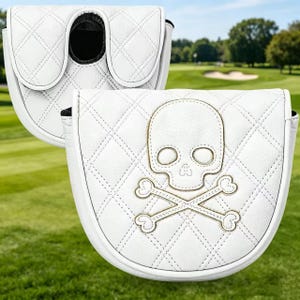 Puede incluir: Una funda para putter de golf blanca con un patr&oacute;n de diamantes acolchado y un dise&ntilde;o bordado de calavera y tibias cruzadas en hilo dorado. La funda tiene una forma redondeada con un mecanismo de cierre seguro visible en la parte superior.