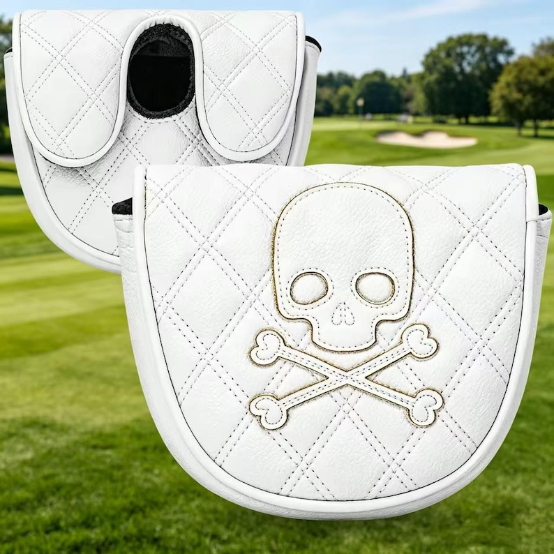 Puede incluir: Una funda para putter de golf blanca con un patr&oacute;n de diamantes acolchado y un dise&ntilde;o bordado de calavera y tibias cruzadas en hilo dorado. La funda tiene una forma redondeada con un mecanismo de cierre seguro visible en la parte superior.