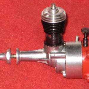 Puede incluir: Un motor de bujía incandescente para aeromodelismo en colores plata y negro, con culata metálica, eje de transmisión horizontal con tuerca para hélice y placa trasera de plástico rojo con un tornillo de ajuste negro en la parte superior.