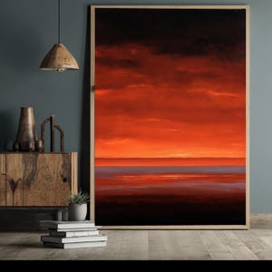 Op de afbeelding: Een ingelijste kunstdruk met een levendige zonsondergang. Het kunstwerk toont een dramatische lucht met tinten rood, oranje en zwart, die weerspiegeld worden in een donker, kalm water. De print wordt getoond in een houten lijst.