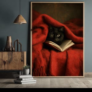 Könnte beinhalten: Gerahmter Druck mit einer schwarzen Katze unter einer roten Decke, die ein Buch liest. Die gelben Augen der Katze sind sichtbar. Der Druck befindet sich in einem Holzrahmen und schafft eine gemütliche Atmosphäre.