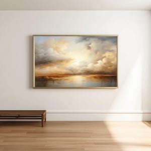 Könnte beinhalten: Gerahmtes Landschaftsgemälde, das einen dramatischen Himmel mit goldenen und grauen Wolken über einem ruhigen Gewässer zeigt. Das Kunstwerk befindet sich in einem goldenen Rahmen und hängt an einer weißen Wand. Darunter befindet sich eine Holzbank.