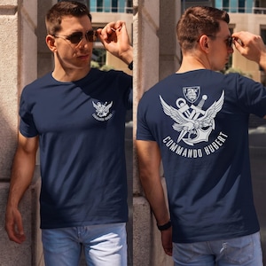 May include: A navy blue short-sleeved t-shirt featuring a white graphic print. The front chest displays a small emblem, while the back showcases a large design of an eagle, anchor, and sword with the text COMMANDO HUBERT printed below.