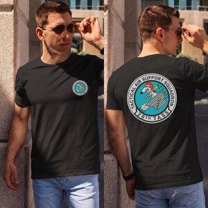 May include: A black short-sleeved t-shirt featuring a circular graphic on the left chest and a larger version on the back. The graphic depicts a cartoon character in a red helmet riding a gray aircraft with the text Tactical Air Support Squadron 20th TASS.