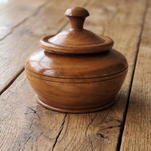 May include: A small, round, polished wooden lidded container with a smooth, warm brown finish. The vessel features a slightly tapered base, a rounded body, and a matching lid topped with a small, knob-like handle centered on the top surface.
