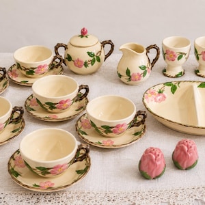 May include: A ceramic tea set featuring a cream-colored base with hand-painted pink wild roses and green leaves. The set includes four teacups with matching saucers, a lidded sugar bowl, a creamer, three egg cups, a divided oval dish, and two salt and pepper shakers shaped like rosebuds.