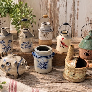 May include: A collection of ceramic decorative items including several small jugs with wire handles, a pig-shaped coin bank with blue floral accents, a numbered crock, and a small watering can, all featuring various painted designs and textures.