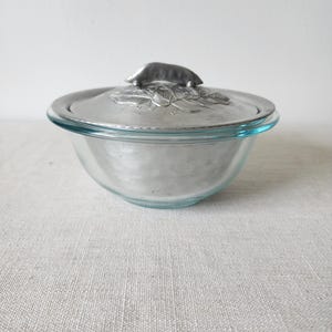 May include: A clear glass bowl with a silver lid. The lid features a raised pea pod design with leaves. The bowl is sitting on a light-colored surface. The bowl is likely used for serving or storage.