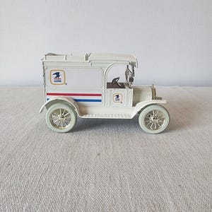 May include: A white vintage U.S. Mail truck model with red and blue stripes. The model features gold-colored wheels and the U.S. Mail logo on the side. The truck has a detailed interior and a roof over the driver's seat.