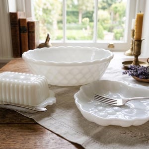 May include: A collection of white milk glass tableware including a large scalloped bowl with a diamond pattern, a rectangular butter dish with vertical ribbing, and a small plate featuring a molded grape cluster design with a silver fork.