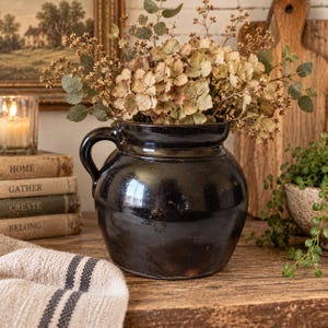 May include: A dark brown, glazed ceramic crock with a single side handle, filled with a bouquet of dried beige hydrangea flowers and green eucalyptus stems. The vessel has a rounded body and a wide, open mouth, showcasing a smooth, reflective finish.