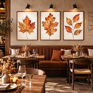 Puede incluir: Tres cuadros de pared con ilustraciones en acuarela de hojas de otoño en tonos naranja, rojo y marrón. Las obras de arte se exhiben sobre un banco de cuero en un comedor con el texto Good Food Brings People Together.