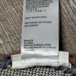 May include: A white fabric label with black text, including care instructions and an address. The label reads "EXCLUSIVE OF BACKING" in several languages. The label is attached to a patterned textile in shades of grey, blue, and brown.