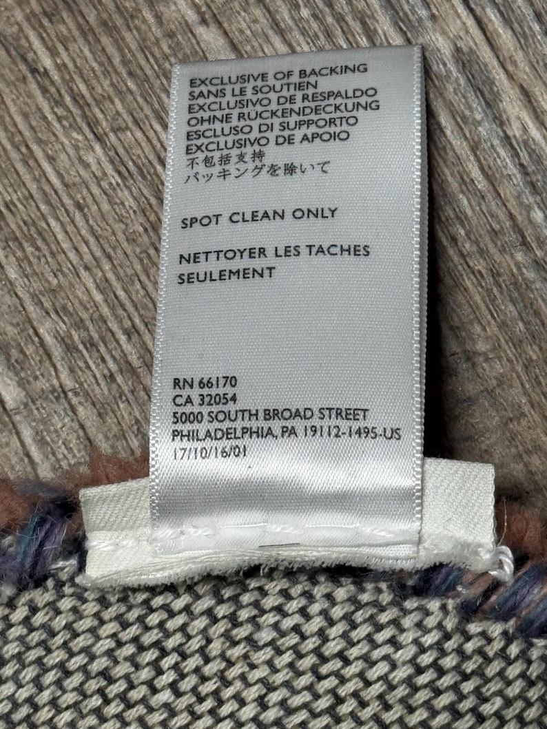 May include: A white fabric label with black text, including care instructions and an address. The label reads "EXCLUSIVE OF BACKING" in several languages. The label is attached to a patterned textile in shades of grey, blue, and brown.