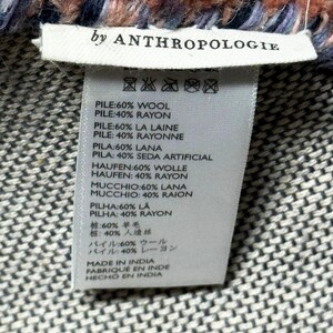 May include: Close-up of a woven textile with a label that reads "by ANTHROPOLOGIE." The textile has a textured, patterned design in shades of grey, with a fluffy, fringed edge in coral, blue, and purple.