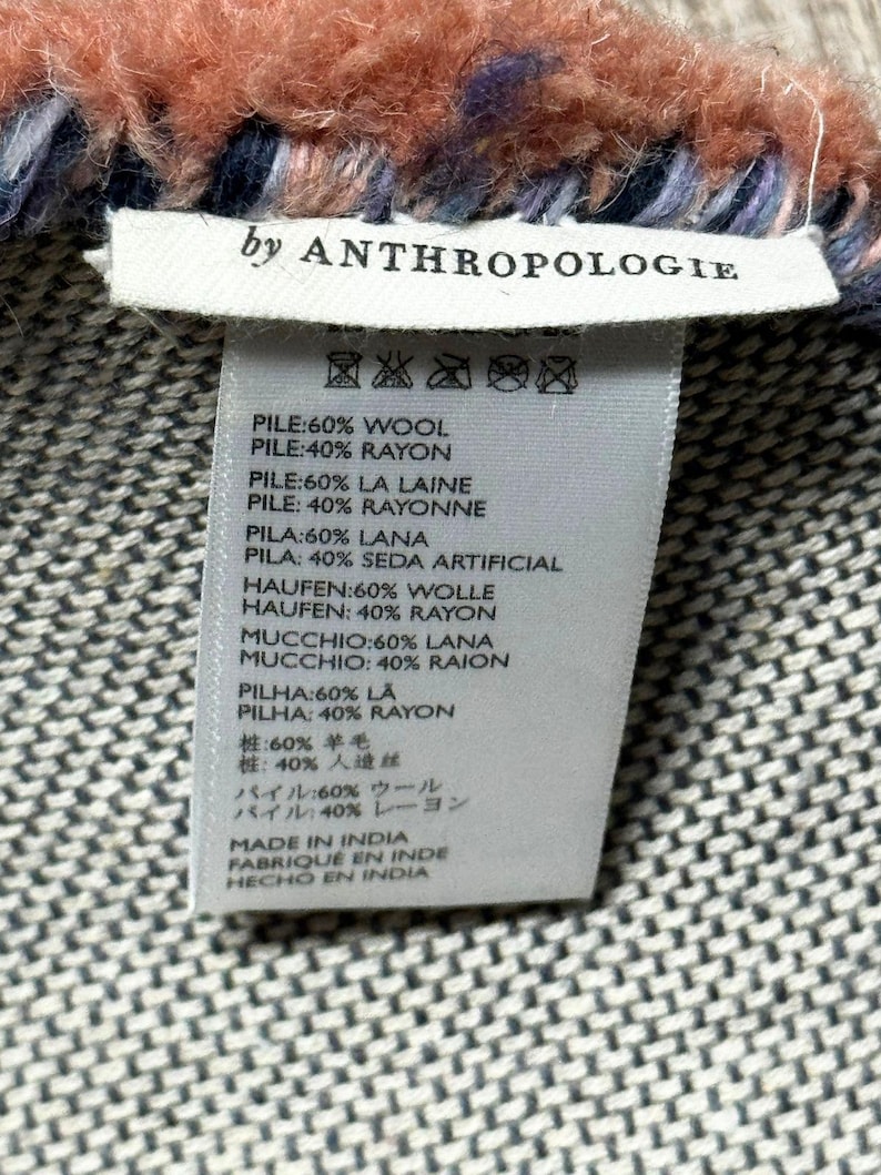 May include: Close-up of a woven textile with a label that reads "by ANTHROPOLOGIE." The textile has a textured, patterned design in shades of grey, with a fluffy, fringed edge in coral, blue, and purple.