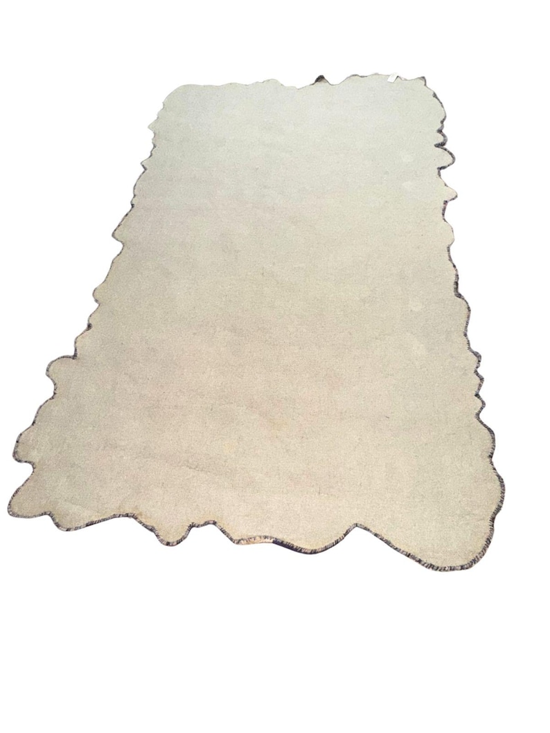 May include: A beige, irregularly shaped rug with a dark border. The rug has a unique, organic form, and the edges are slightly frayed, giving it a rustic appearance. The rug is set against a white background.