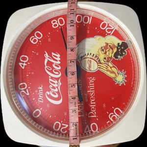 May include: A white analog scale with a red circular face featuring Coca-Cola branding, a vintage illustration of a person, and the text Drink Coca-Cola and Delicious and Refreshing. A measuring tape shows the scale face is approximately 11 inches wide.