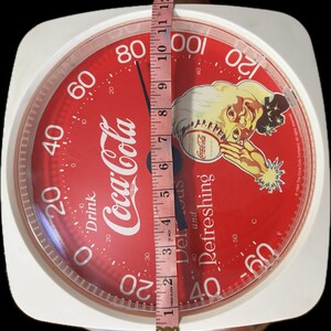 May include: A round Coca-Cola branded kitchen scale with a red face, white numbers, and a graphic of a person. A pink measuring tape is placed vertically across the center, showing the scale has a diameter of approximately 12 inches.