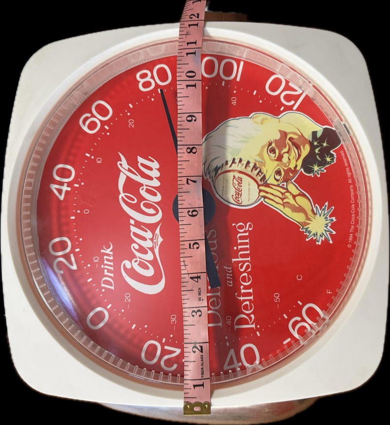 May include: A round Coca-Cola branded kitchen scale with a red face, white numbers, and a graphic of a person. A pink measuring tape is placed vertically across the center, showing the scale has a diameter of approximately 12 inches.