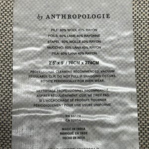 Puede incluir: Una etiqueta rectangular de Anthropologie, con texto que detalla la composici&oacute;n de la tela (60% lana, 40% ray&oacute;n), las dimensiones (2'5" x 9" / 76cm x 274cm) y las instrucciones de cuidado. La etiqueta tambi&eacute;n incluye los n&uacute;meros RN y CA, y est&aacute; hecha en India.
