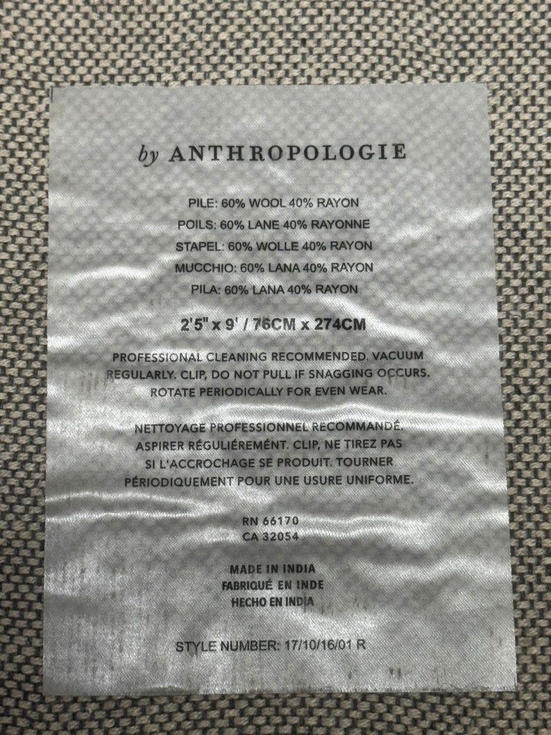 Puede incluir: Una etiqueta rectangular de Anthropologie, con texto que detalla la composici&oacute;n de la tela (60% lana, 40% ray&oacute;n), las dimensiones (2'5" x 9" / 76cm x 274cm) y las instrucciones de cuidado. La etiqueta tambi&eacute;n incluye los n&uacute;meros RN y CA, y est&aacute; hecha en India.