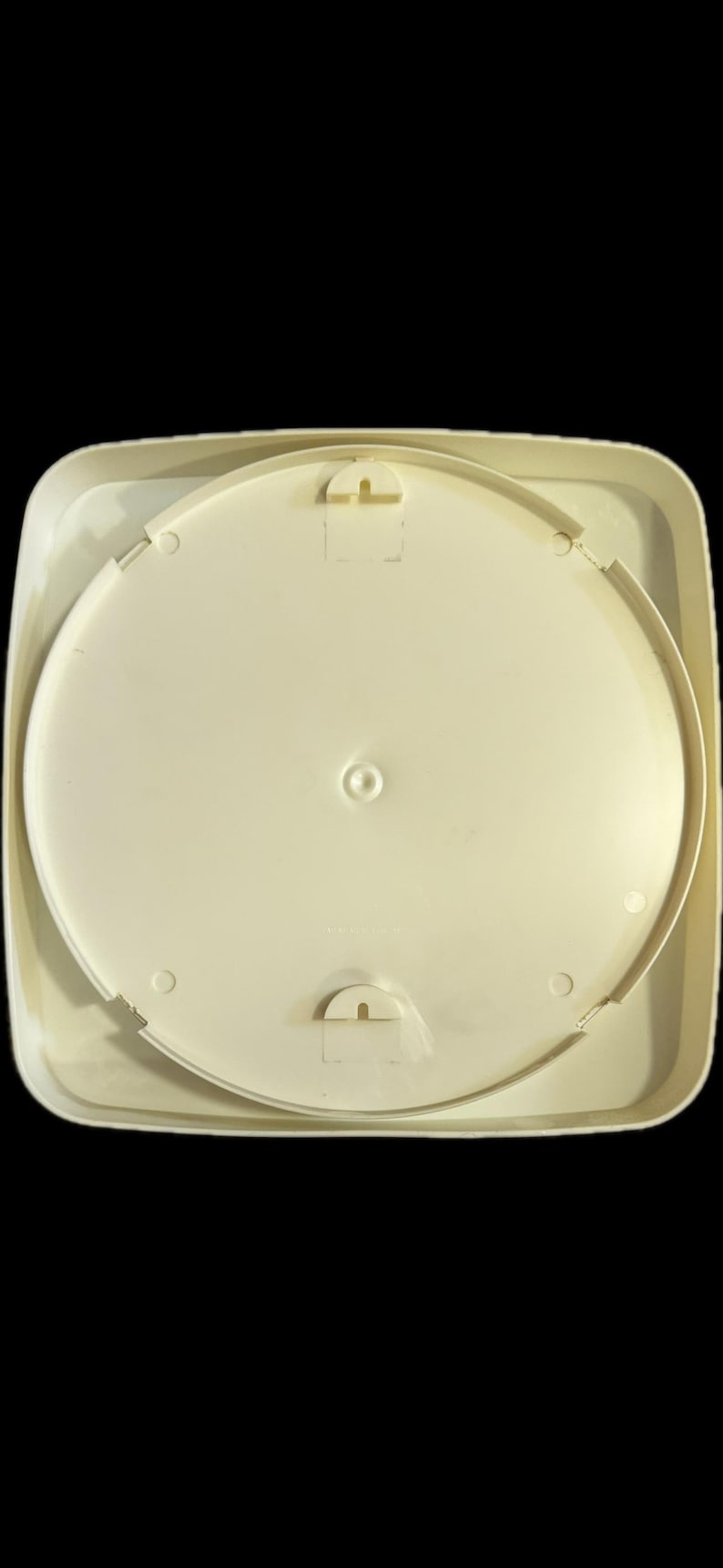 May include: A square, off-white plastic ceiling light fixture base with rounded corners. The center features a circular mounting plate with a central screw hole and two semi-circular locking tabs positioned at the top and bottom for secure installation.