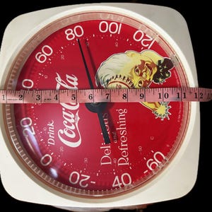May include: A square white Coca-Cola thermometer with a red circular face featuring a vintage illustration of a person, the text Drink Coca-Cola Delicious and Refreshing, and a measuring tape showing a width of approximately 13 inches.