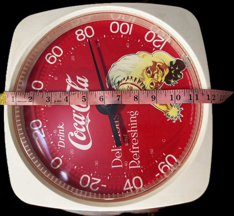 May include: A square white Coca-Cola thermometer with a red circular face featuring a vintage illustration of a person, the text Drink Coca-Cola Delicious and Refreshing, and a measuring tape showing a width of approximately 13 inches.