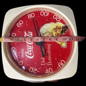 May include: A square Coca-Cola branded kitchen scale with a red face, white text, and a graphic of a person. A measuring tape across the center shows the scale is approximately 11 inches wide. The dial features numbers from -60 to 120.
