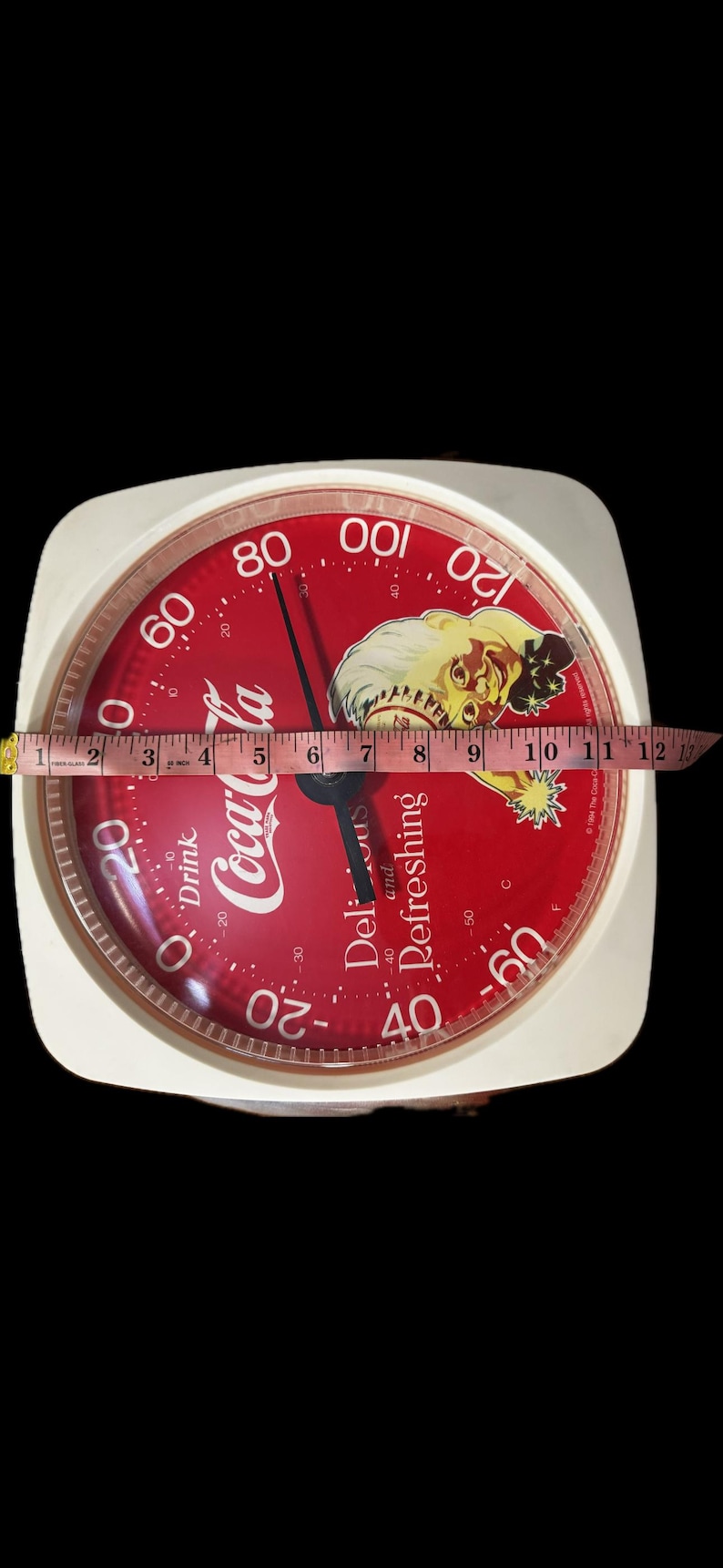 May include: A square Coca-Cola branded kitchen scale with a red face, white text, and a graphic of a person. A measuring tape across the center shows the scale is approximately 11 inches wide. The dial features numbers from -60 to 120.