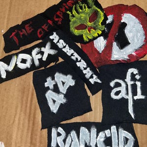 May include: A collection of black fabric patches featuring hand-painted white and yellow designs. The patches display band names and logos including The Offspring, NOFX, AFI, Rancid, 4+4, and a skull graphic, along with a red and white circular symbol.