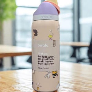 Może przedstawiać: Beżowa butelka na wodę Owala z fioletową pokrywką i wbudowaną słomką. Butelka zawiera ilustracje postaci z kresk&oacute;wek oraz tekst "Hi there. I'm leak proof. I'm insulated. And I have a built-in straw." Pojemność 945 ml.