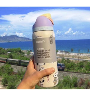 K&ouml;nnte beinhalten: Eine cremefarbene Owala-Trinkflasche mit Snoopy-Design und lila und gelbem Deckel. Die Flasche hat einen eingebauten Strohhalm und den Text: "Hi there. I'm leak proof. I'm insulated. And I have a built-in straw."