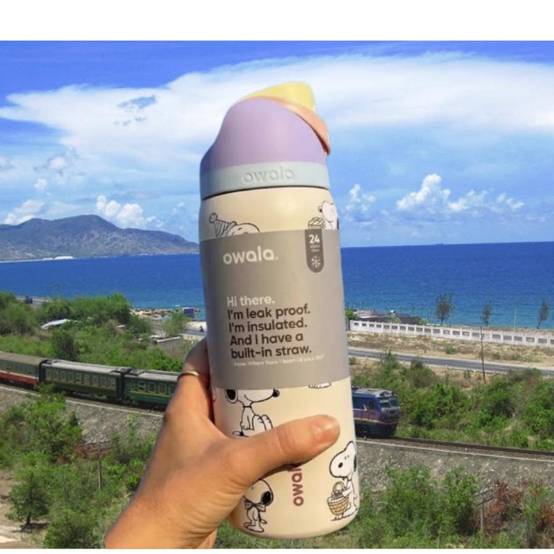 K&ouml;nnte beinhalten: Eine cremefarbene Owala-Trinkflasche mit Snoopy-Design und lila und gelbem Deckel. Die Flasche hat einen eingebauten Strohhalm und den Text: "Hi there. I'm leak proof. I'm insulated. And I have a built-in straw."