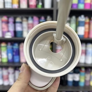 K&ouml;nnte beinhalten: Nahaufnahme eines hellbeigen Becherdeckels mit schwarzer Gummidichtung. Der Deckel hat eine kreisf&ouml;rmige &Ouml;ffnung und einen Strohhalmmechanismus. Im Hintergrund sind Regale mit verschiedenen Flaschen zu sehen.