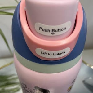 K&ouml;nnte beinhalten: Eine rosa-blaue Wasserflasche mit einem Druckknopfdeckel. Der Deckel ist hellrosa mit einem blauen und korallenfarbenen Akzent. Die W&ouml;rter "Push Button" und "Lift to Unlock" sind auf dem Deckel aufgedruckt. Die Flasche hat Cartoon-Grafiken.