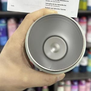 K&ouml;nnte beinhalten: Ein hellgrauer isolierter Becher mit silbernem Rand, gehalten in einer Hand. Der Innenraum ist dunkler grau. Ein Papier mit Text in mehreren Sprachen wird &uuml;ber dem Becher gehalten. Der Hintergrund zeigt Regale mit bunten Flaschen.