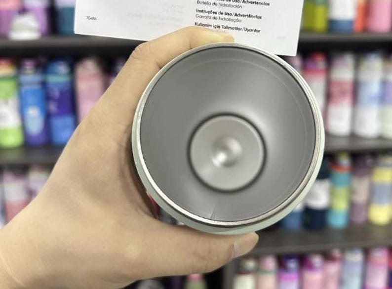 K&ouml;nnte beinhalten: Ein hellgrauer isolierter Becher mit silbernem Rand, gehalten in einer Hand. Der Innenraum ist dunkler grau. Ein Papier mit Text in mehreren Sprachen wird &uuml;ber dem Becher gehalten. Der Hintergrund zeigt Regale mit bunten Flaschen.