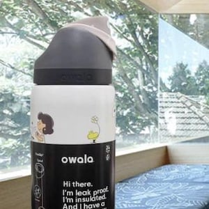 K&ouml;nnte beinhalten: Owala Isolierflasche in Wei&szlig; und Schwarz mit eingebautem Strohhalm. Die Flasche hat einen schwarzen Deckel und einen wei&szlig;en K&ouml;rper mit Cartoon-Illustrationen und dem Text "Hi there. I'm leak proof. I'm insulated. And I have a built-in straw. 945ml."