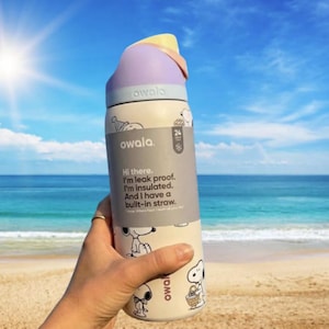 May include: A cream-colored Owala water bottle with a Snoopy cartoon design, a purple lid, and a yellow accent. The bottle has a built-in straw and text that reads "Hi there. I'm leak proof. I'm insulated. And I have a built-in straw."