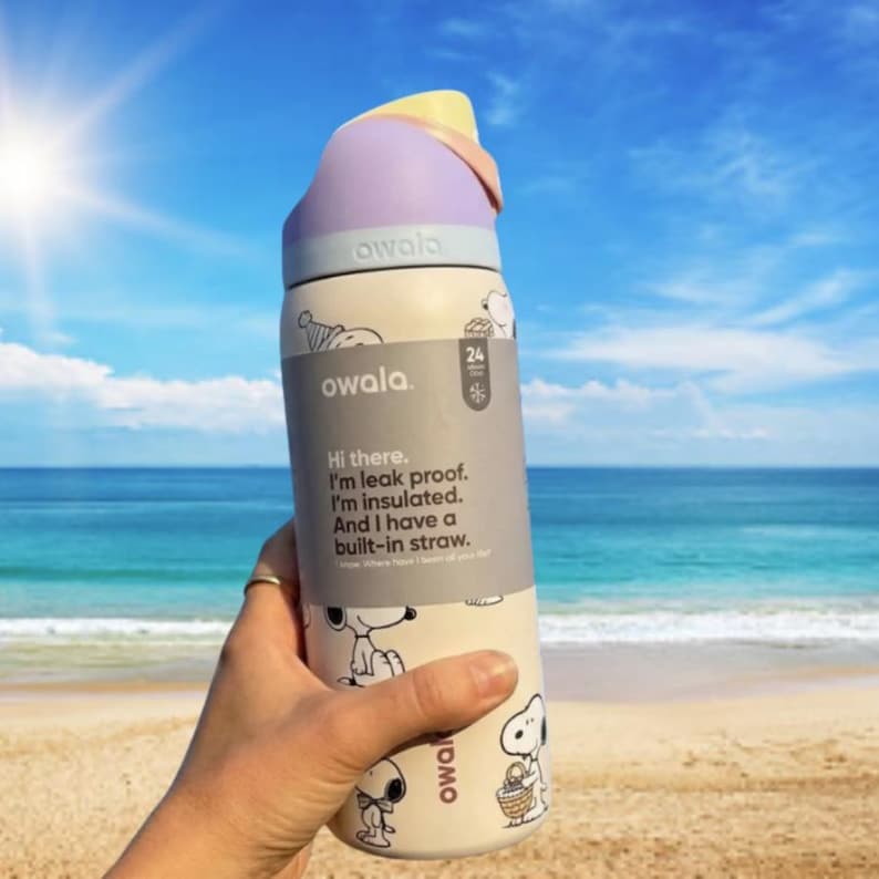 May include: A cream-colored Owala water bottle with a Snoopy cartoon design, a purple lid, and a yellow accent. The bottle has a built-in straw and text that reads "Hi there. I'm leak proof. I'm insulated. And I have a built-in straw."